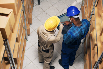 Diverse employees analyzing inventory list on laptop, counting products in boxes on storage room shelves. Young people using device to work on stock logistics, delivery service.